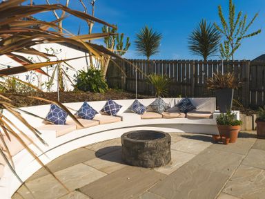 An outdoor patio with a curved bench with cushions and pillows around a stone fire pit at Oystercatcher in Mevagissey