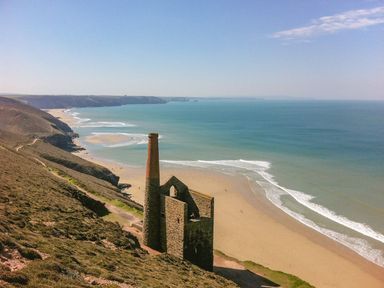 An old stone building with a tall chimney on a cliff overlooking a sandy beach and ocean waves at 2 Rose Cottage in St Agnes