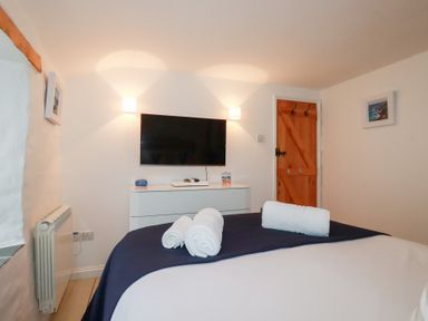 A bedroom with a bed with rolled towels a wall mounted tv a white dresser a wooden door and framed pictures at 2 Rose Cottage in St Agnes