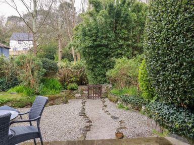 A garden area with a gravel pathway leading to a wooden gate surrounded by bushes and trees at 2 Rose Cottage in St Agnes