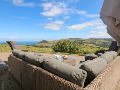 An outdoor seating area with wicker furniture and cushions overlooking fields and hills at Buddicombe House in Combe Martin