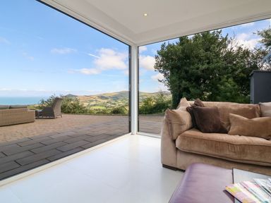 A corner of a living room with a sofa and glass walls showing an outdoor patio and countryside view at Buddicombe House in Combe Martin