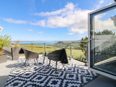 A balcony with two chairs a small table and a patterned rug overlooking fields and ocean at Buddicombe House in Combe Martin