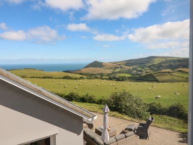 A patio with outdoor seating and umbrella overlooking fields with sheep and hills at Buddicombe House in Combe Martin