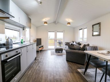 An open plan kitchen and living area with gray cupboards and oven a sofa a dining table two chairs and a television at Cuckoo in Ramsey Huntingdon