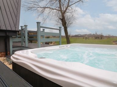 An outdoor hot tub next to a wooden deck and railing with a tree and open field in the background at Sunflower Lodge Near Misterton