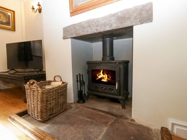 A room with a wood burning stove in a stone hearth with a basket of firewood and a television on a wooden table at Court House Cottage in Gatcombe near Blakeney