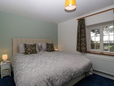 A bedroom with a large bed with patterned pillows bedside tables lamps a window with patterned curtains and a radiator at Court House Cottage in Gatcombe near Blakeney