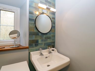 A bathroom sink with two taps a round mirror and a soap dispenser by a window at Court House Cottage in Gatcombe near Blakeney