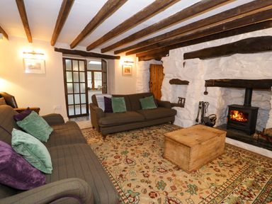 A living room with two sofas, a wooden chest coffee table, a patterned rug, a wood-burning stove, and exposed wooden beams at Glandwr Cottage in Llanbedr