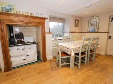 A dining area with a wooden table and six chairs next to a vintage white stove and a window at Glandwr Cottage in Llanbedr