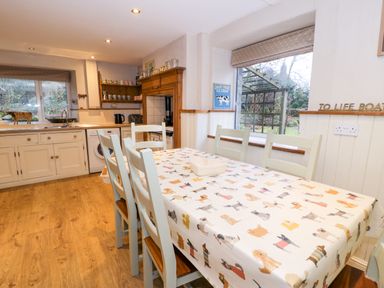 A kitchen with a dining table covered with a dog patterned tablecloth and wooden chairs at Glandwr Cottage in Llanbedr