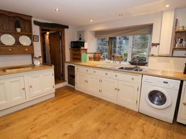 A kitchen with white cabinets a wooden floor a washing machine and a window showing a stone wall at Glandwr Cottage in Llanbedr