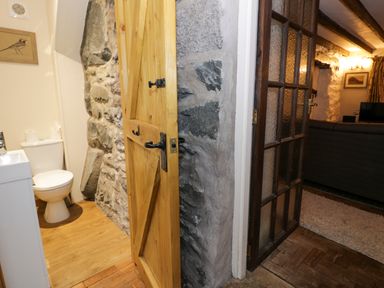 A bathroom with a toilet and sink visible through a wooden door and a living room with a dark sofa and television behind a glass paneled door at Glandwr Cottage in Llanbedr