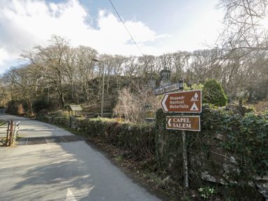 A road with signs pointing to Rhaeadr Nantcol Waterfalls and Capel Salem with trees and stone walls at Glandwr Cottage in Llanbedr