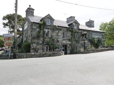 A stone building with climbing plants and multiple chimneys by a roadside with directional signs at Glandwr Cottage in Llanbedr