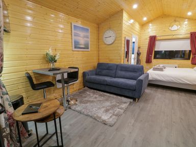 A wooden cabin interior with a sofa a bed a dining table with two chairs and a wall clock at Binevanagh Cabin in Magilligan near Castlerock
