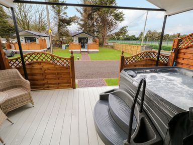A covered porch with a hot tub and wicker chair overlooking a lawn and small cabins at Binevanagh Cabin in Magilligan near Castlerock