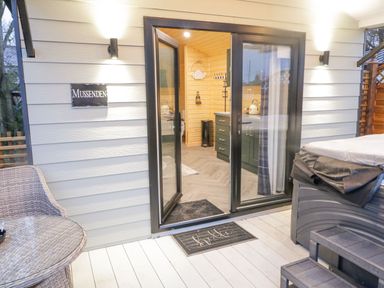 An open glass door leading to a kitchenette with cabinets and a kettle outside on a wooden deck with a wicker chair table and a hot tub at MUSSENDEN CABIN in Magilligan near Castlerock