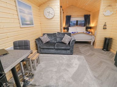 A wooden cabin interior with a grey sofa and table and stools and a bed in the background with curtains at MUSSENDEN CABIN Magilligan near Castlerock