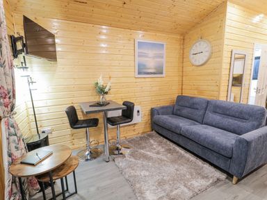 A living area with a blue sofa a small table with two chairs a wall clock and a mounted television at Benone Cabin Magilligan near Castlerock
