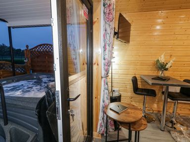 A wooden interior room with a small round table and tall chairs next to a mounted television and a hot tub outside at Benone Cabin in Magilligan near Castlerock