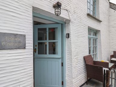 A blue door on a white textured wall with a sign reading Teana Cottage and wicker chairs outside at Teana Cottage in Polperro