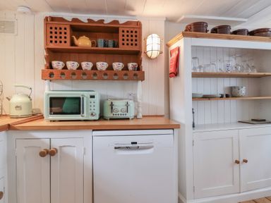 A kitchen countertop with a microwave, toaster, and kettle under a wooden shelf with cups and teapots at Teana Cottage in Polperro