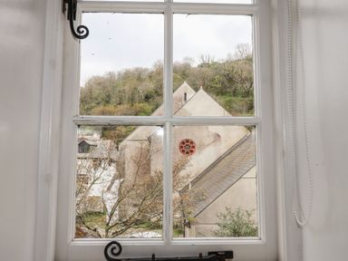 View through a window showing a building with a triangular roof and circular window near a hillside with trees at Teana Cottage in Polperro