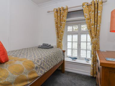 A bedroom with a single bed next to a window with yellow patterned curtains and a wooden dresser with an orange lamp at Teana Cottage in Polperro