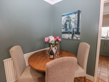 A dining area with a round wooden table surrounded by four beige upholstered chairs a bottle and four glasses on the table a vase of flowers in the corner and a painting on a green wall at Greenhaven in Narberth