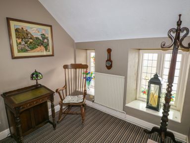 A small room with striped carpet a wooden rocking chair an antique side table with a lamp framed painting on the wall stained glass windows clock and a standing coat rack at Greenhaven in Narberth
