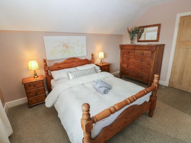 A bedroom with a wooden bed frame two bedside tables with lamps a wooden dresser with a mirror and flowers on top at Greenhaven in Narberth