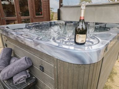 An outdoor hot tub with a bottle of prosecco and three glasses on the edge near rolled towels at Greenhaven in Narberth