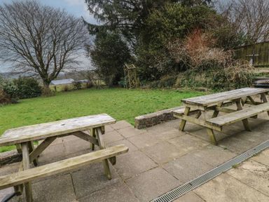 An outdoor area with wooden picnic tables on a paved patio and a grass lawn with trees and bushes at Greenhaven in Narberth