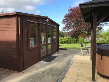 A wooden shed with glass doors on a patio next to outdoor chairs and a covered hot tub with trees and grass in the background at Greenhaven in Narberth