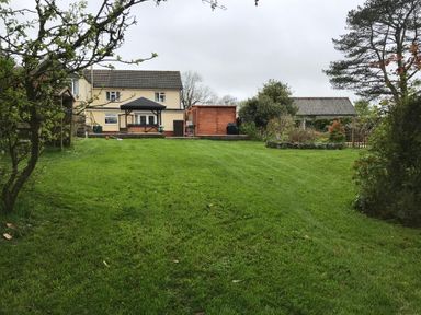 A backyard with a large lawn, a two-story house, a wooden shed, trees, and garden beds at Greenhaven in Narberth