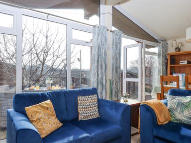 A living room with blue sofas and patterned cushions next to large windows and glass doors at Matilda Lodge in Newton Ferrers