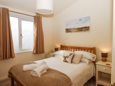 A bedroom with a wooden bed frame white bedding two bedside tables with lamps and a window with curtains at Matilda Lodge in Newton Ferrers
