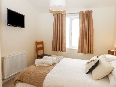 A bedroom with a bed with pillows and towels a wooden chair a window with curtains a radiator and a wall mounted tv at Matilda Lodge in Newton Ferrers