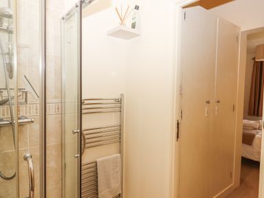 A bathroom with a glass shower enclosure a chrome heated towel rail and a small shelf with toiletries a bedroom is visible through the doorway at Matilda Lodge in Newton Ferrers