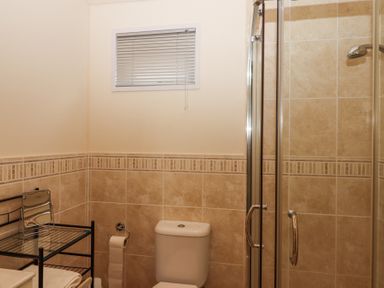 A bathroom with a toilet a glass shower enclosure a window with blinds and a metal rack with towels and a mirror at Matilda Lodge Newton Ferrers
