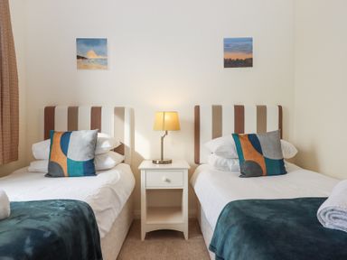 A bedroom with two single beds separated by a white nightstand with a lamp and framed pictures on the wall at Matilda Lodge in Newton Ferrers