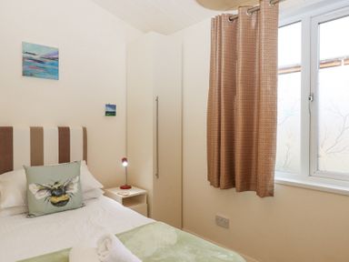 A bedroom with a bed with a bee pillow a white bedside table with a red lamp a tall white wardrobe and a window with beige curtains at Matilda Lodge in Newton Ferrers