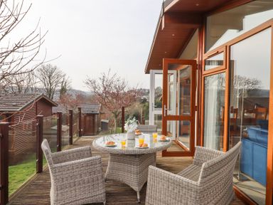 An outdoor wooden deck with a round glass table and four wicker chairs breakfast setup with croissants and orange juice Matilda Lodge Newton Ferrers
