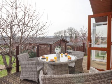An outdoor patio with a round glass table and four wicker chairs with croissants and orange juice at Matilda Lodge in Newton Ferrers