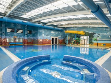 An indoor swimming pool with a circular hot tub a yellow water slide and murals on the walls at Ripples & Reeds - Billing Aquadrome