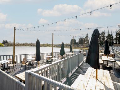 An outdoor seating area with tables, chairs, and closed umbrellas by a body of water at Ripples & Reeds - Billing Aquadrome