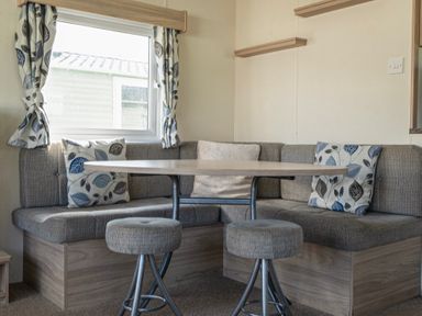 A dining area with a wooden table surrounded by gray cushioned bench seating and two stools at Ripples & Reeds - Billing Aquadrome in Billing Aquadrome Holiday Park