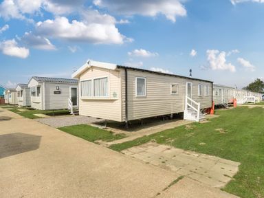 A row of beige and white mobile homes on grassy plots with a paved path and blue sky at Ripples & Reeds - Billing Aquadrome in Billing Aquadrome Holiday Park